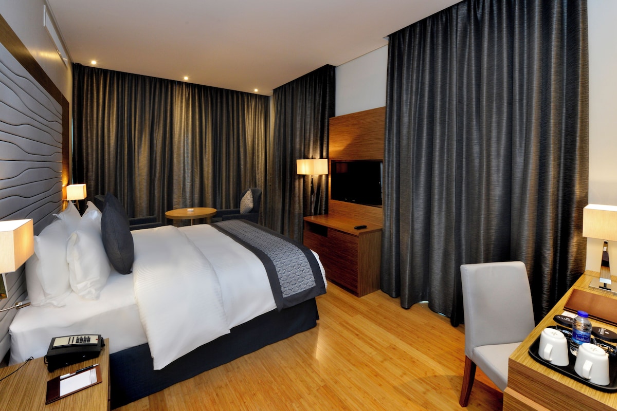 Een hotelkamer met een bed, nachtkastje, lamp, bureau, stoel en een televisie.