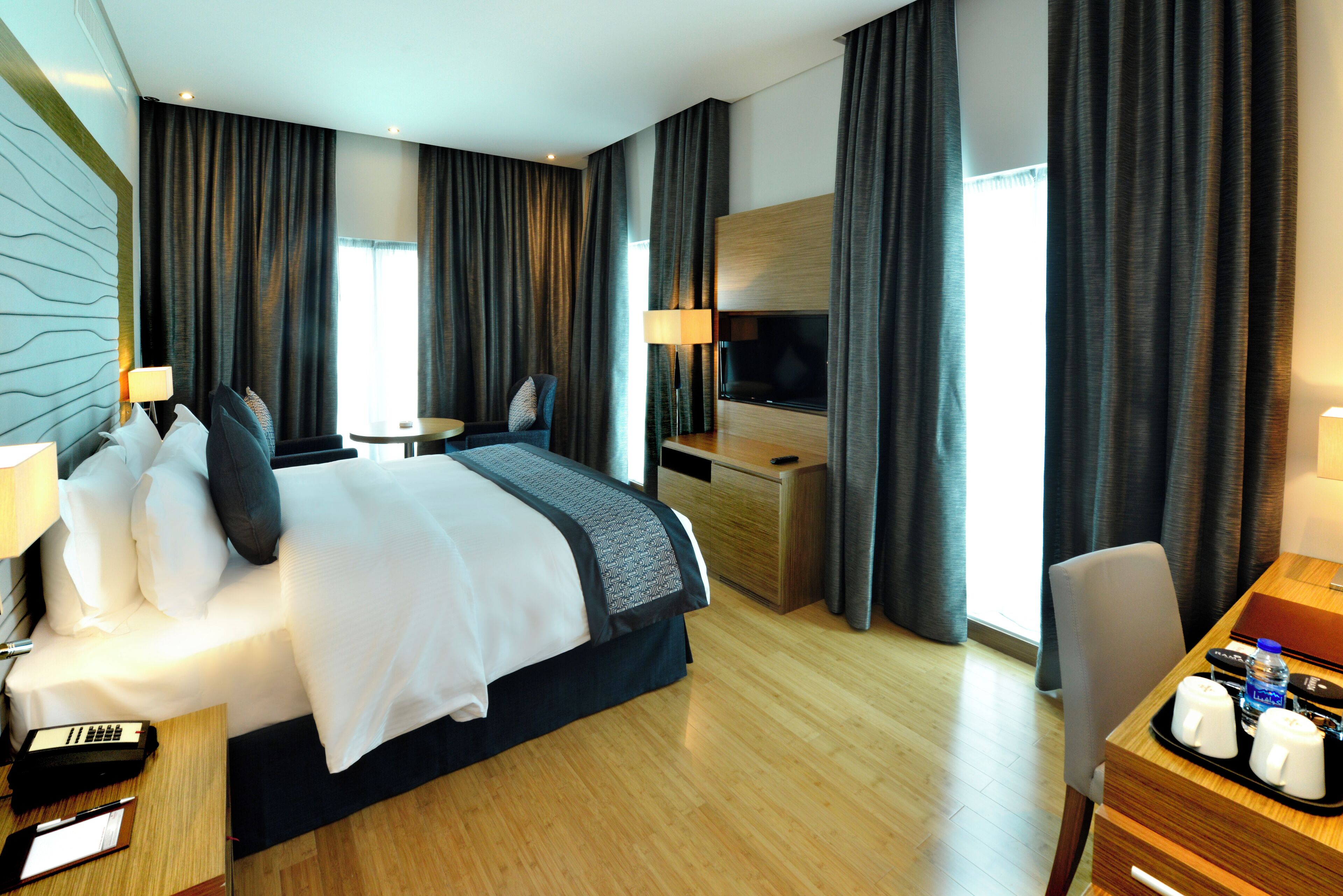 A hotel room with a bed, bedside table, lamp, desk, chair, TV, and curtains.