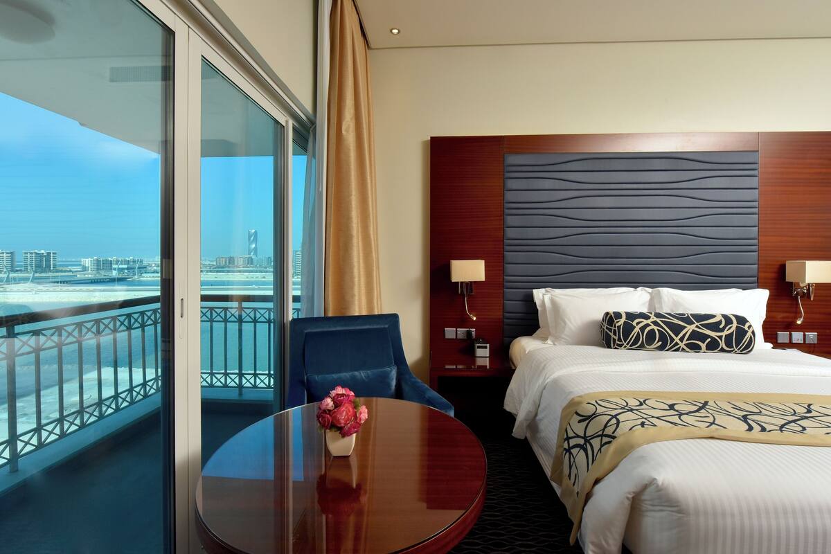 Une chambre d’hôtel avec un grand lit, une table de chevet avec une lampe, une chaise, une table ronde ornée de fleurs et une vue sur la ville et l’eau, grâce à de grandes fenêtres.