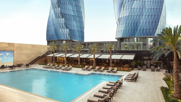 Ein modernes Hotel mit zwei markanten Glasgebäuden, einem Poolbereich mit Liegestühlen und einer Treppe, die zu den oberen Etagen führt.