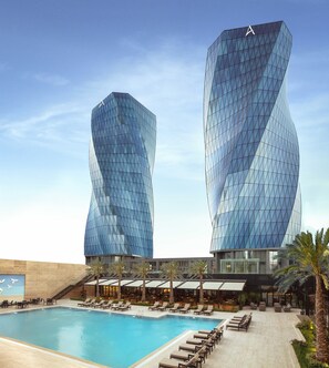 Un hotel moderno con dos edificios de cristal destacados, una zona de piscina con tumbonas y una escalera que lleva a las plantas superiores.