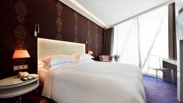 Une chambre d’hôtel avec un grand lit, une table de chevet avec une lampe, une chaise et une grande fenêtre avec des rideaux.