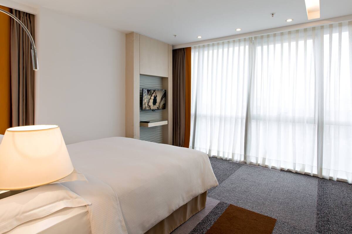 A hotel room with a bed, a desk, a lamp, a TV, and a large window with curtains.