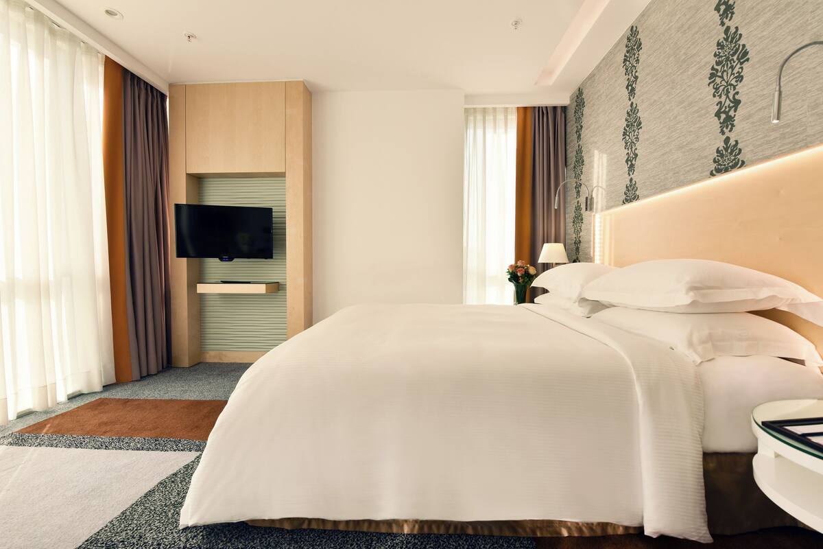 A modern hotel room with a large bed, a flat-screen TV mounted on the wall, a bedside table with a lamp, and a decorative wall panel.