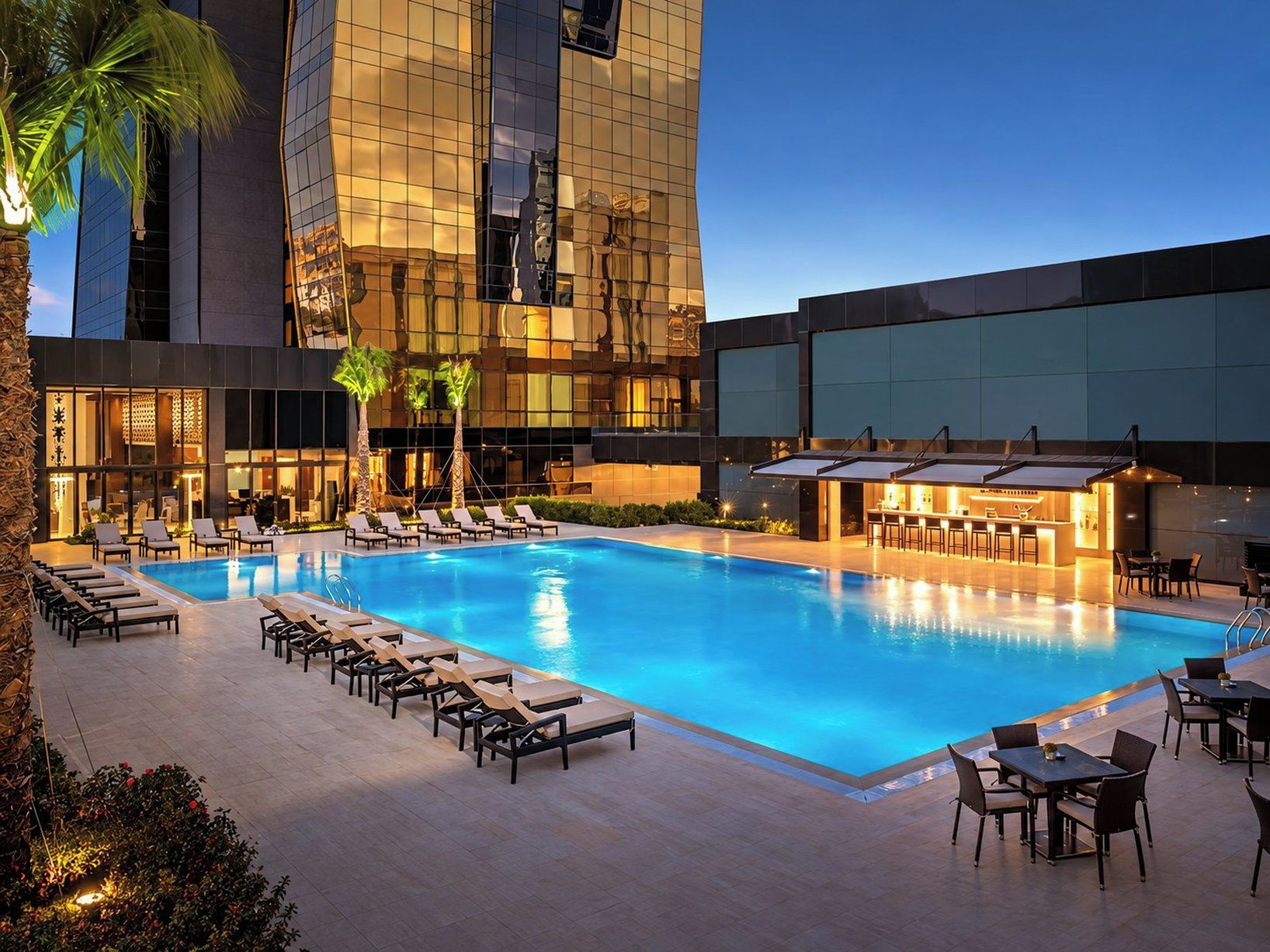 A swimming pool with lounge chairs and tables, surrounded by a modern building with reflective glass windows.