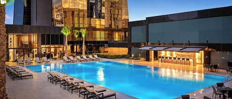 A swimming pool with lounge chairs and tables, surrounded by a modern building with reflective glass windows.
