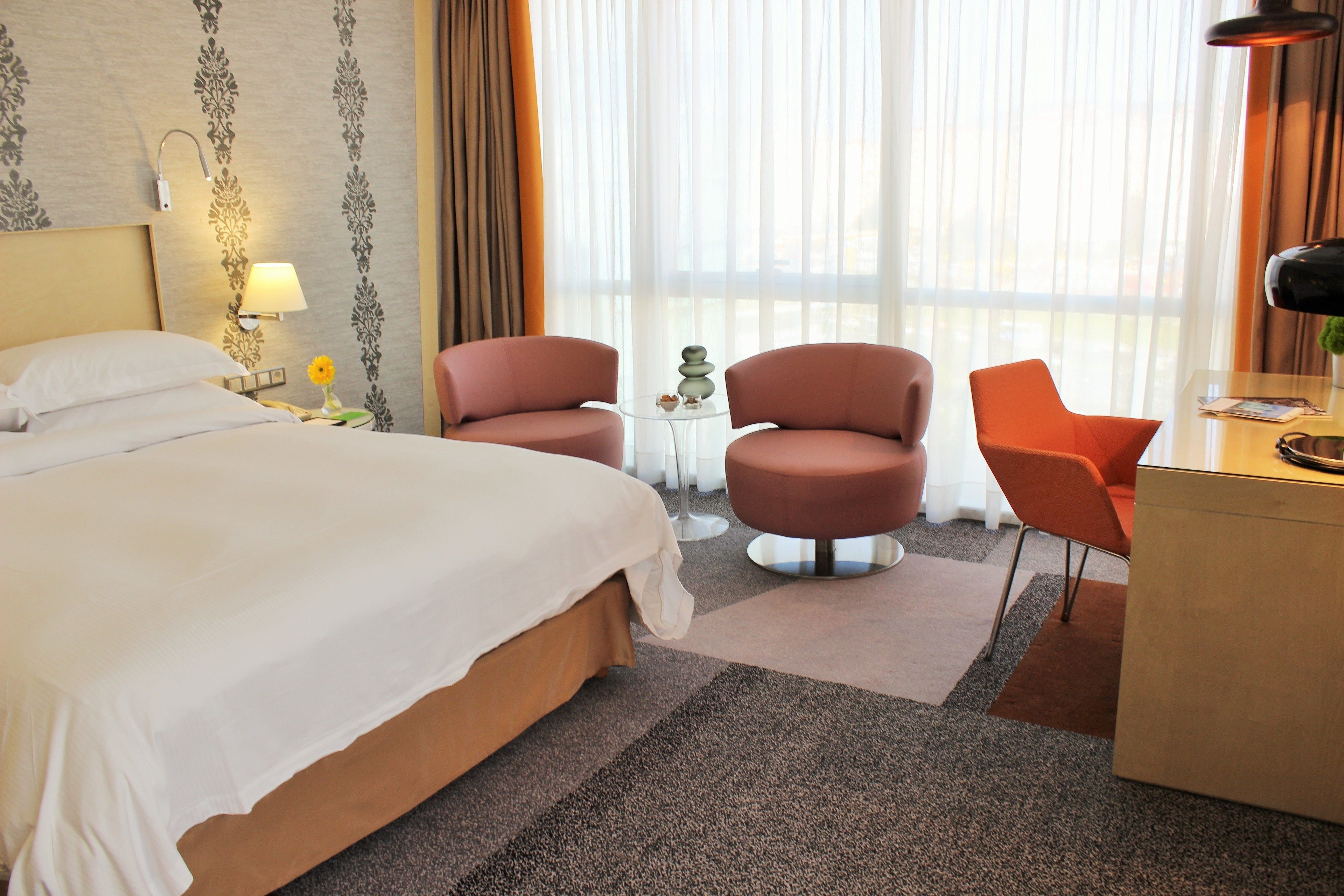 Hotellihuone, jossa on sänky, kaksi nojatuolia, työpöytä, lamppu, maljakko kukkineen ja ikkuna, jossa on läpikuultavat verhot.