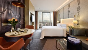 A hotel room with a bed, a dining table with chairs, a coffee table, a view of the city through large windows, and a reception desk. - Tango Arjaan by Rotana Istanbul (Istanbul)