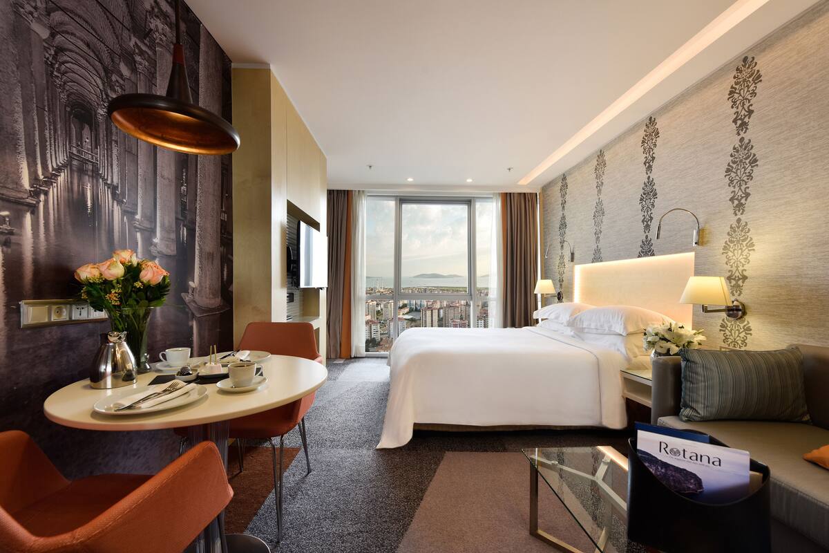 A hotel room with a bed, a dining table with chairs, a coffee table, a view of the city through large windows, and a reception desk.