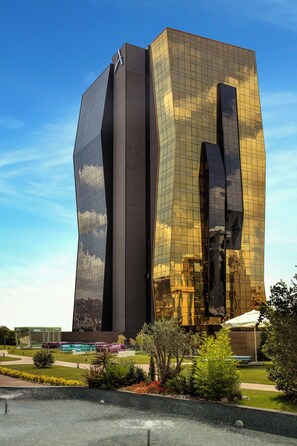 Een modern gebouw met een uniek hoekig ontwerp, een spiegelende glazen gevel en een aangelegde omgeving met zitplaatsen en een zwembad.