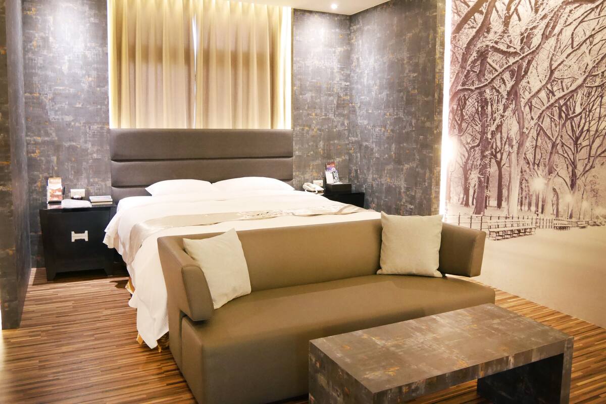 Quarto de hotel moderno com uma cama grande, um sofá, uma mesa de cabeceira e um mural na parede.