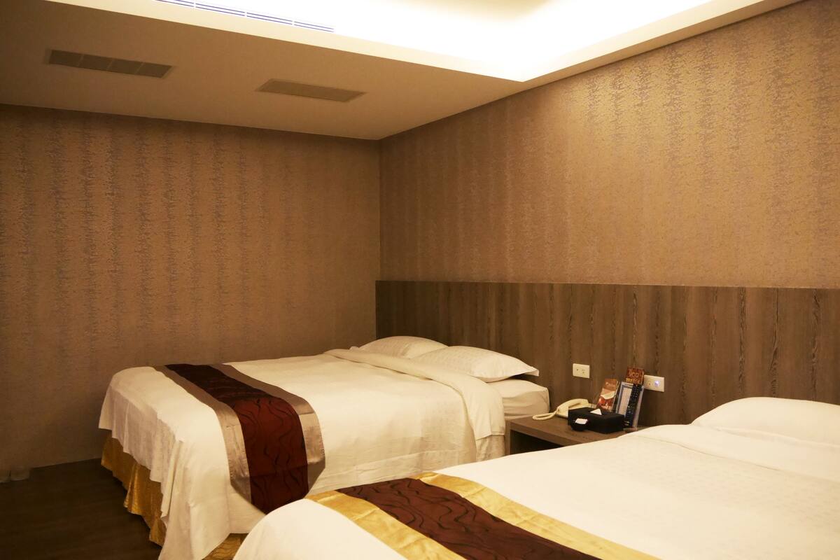 Quarto de hotel com duas camas, uma cabeceira de madeira, mesa de cabeceira com telefone e revista, e luminária fixada na parede.
