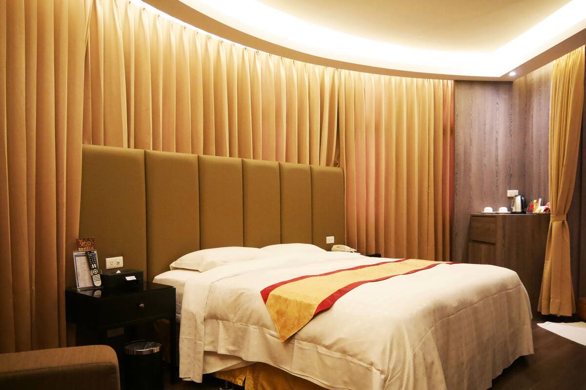 Quarto de hotel com uma cama grande, uma mesinha de cabeceira, um criado-mudo e um guarda-roupa de madeira.