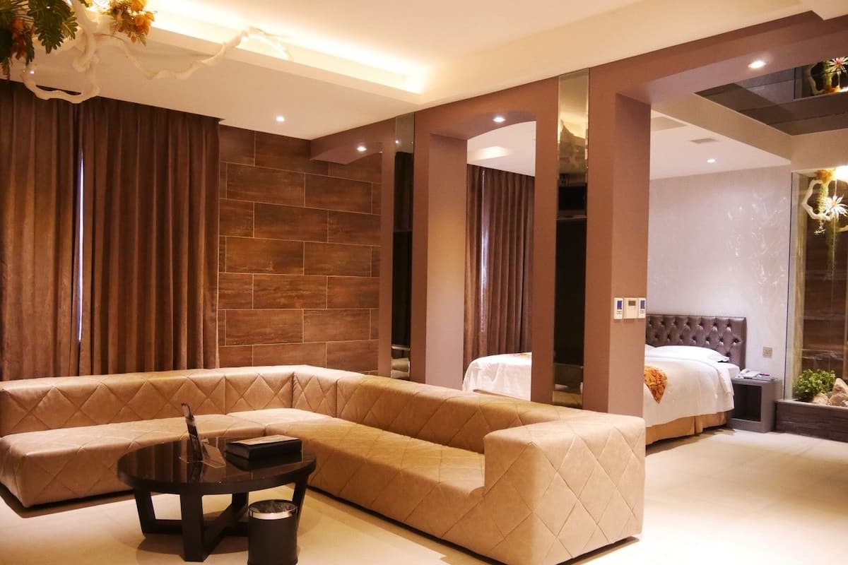 Quarto de hotel moderno com um amplo sofá seccional bege, uma mesa redonda pequena e uma cama com cabeceira estofada.