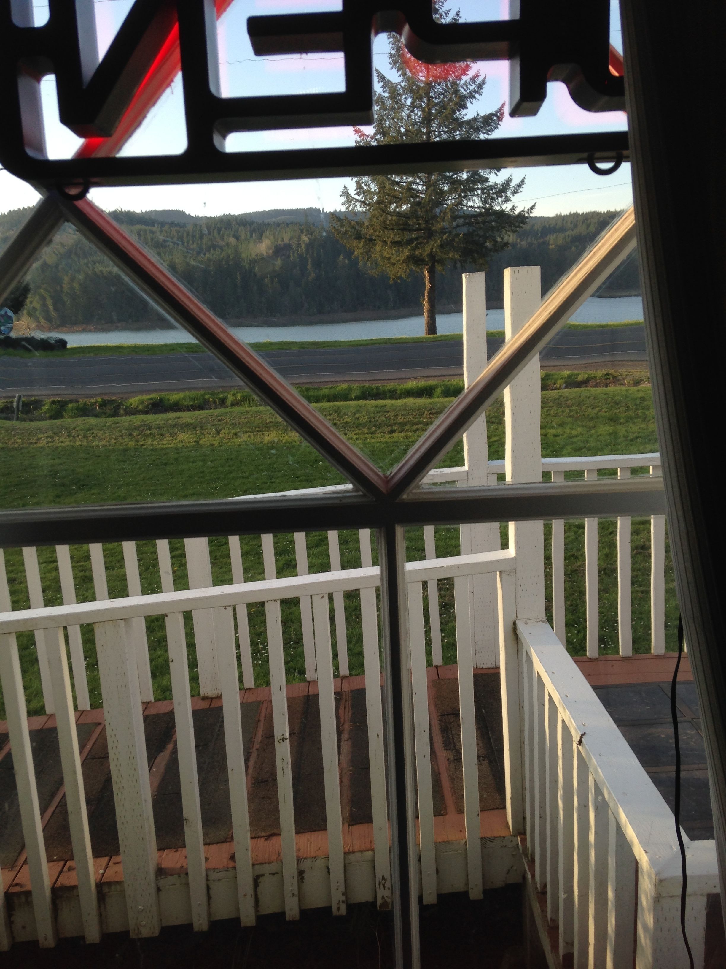A view from a balcony with a white railing, overlooking a lake, a road, and a forested area.