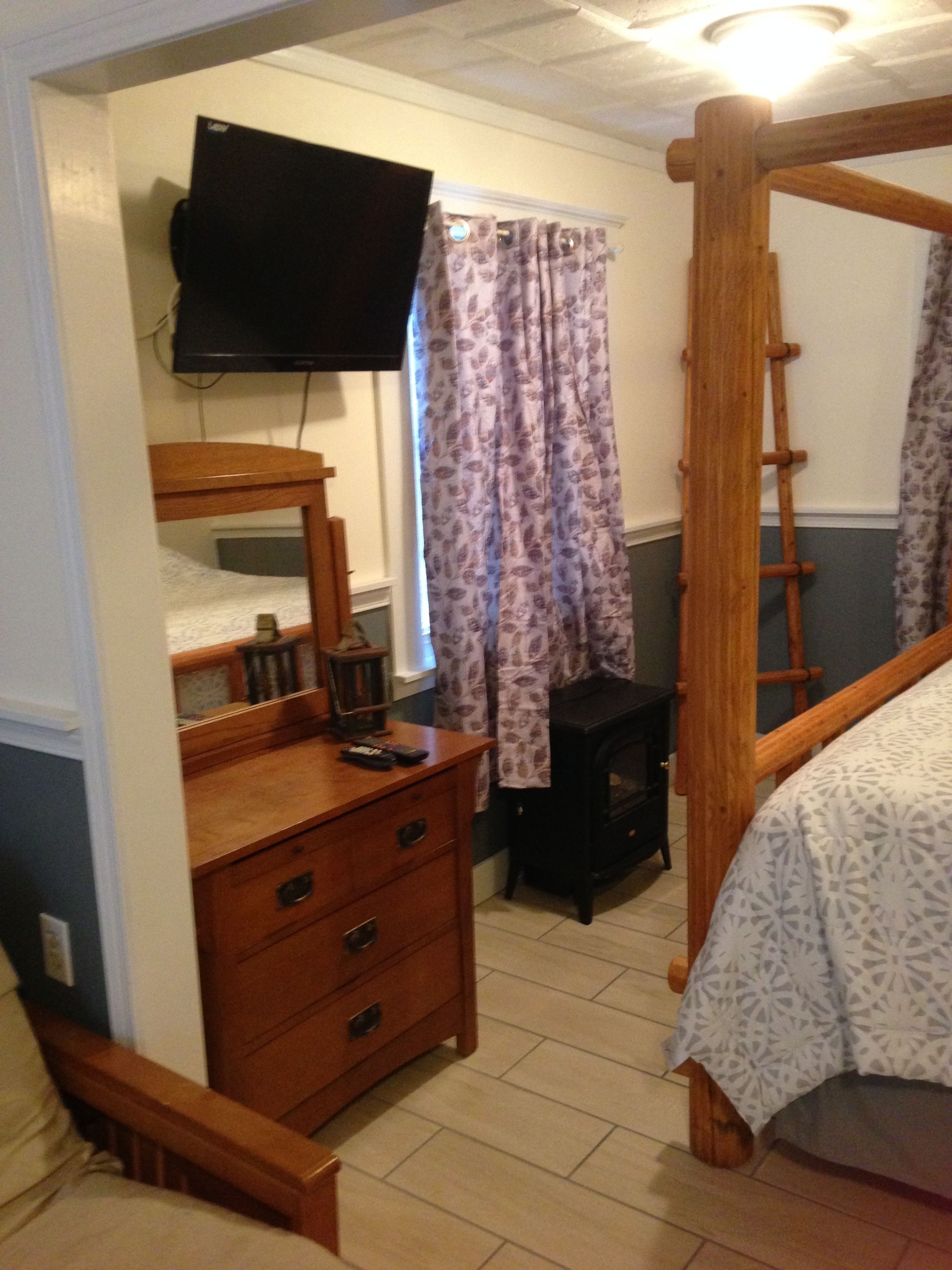 A bedroom with a wooden bed, a dresser, a television, a small black heater, and a window with floral curtains.
