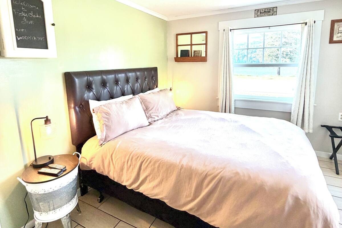 A bedroom with a bed, a bedside table with a lamp, a window with curtains, a wall-mounted sign, and a small window with a view.