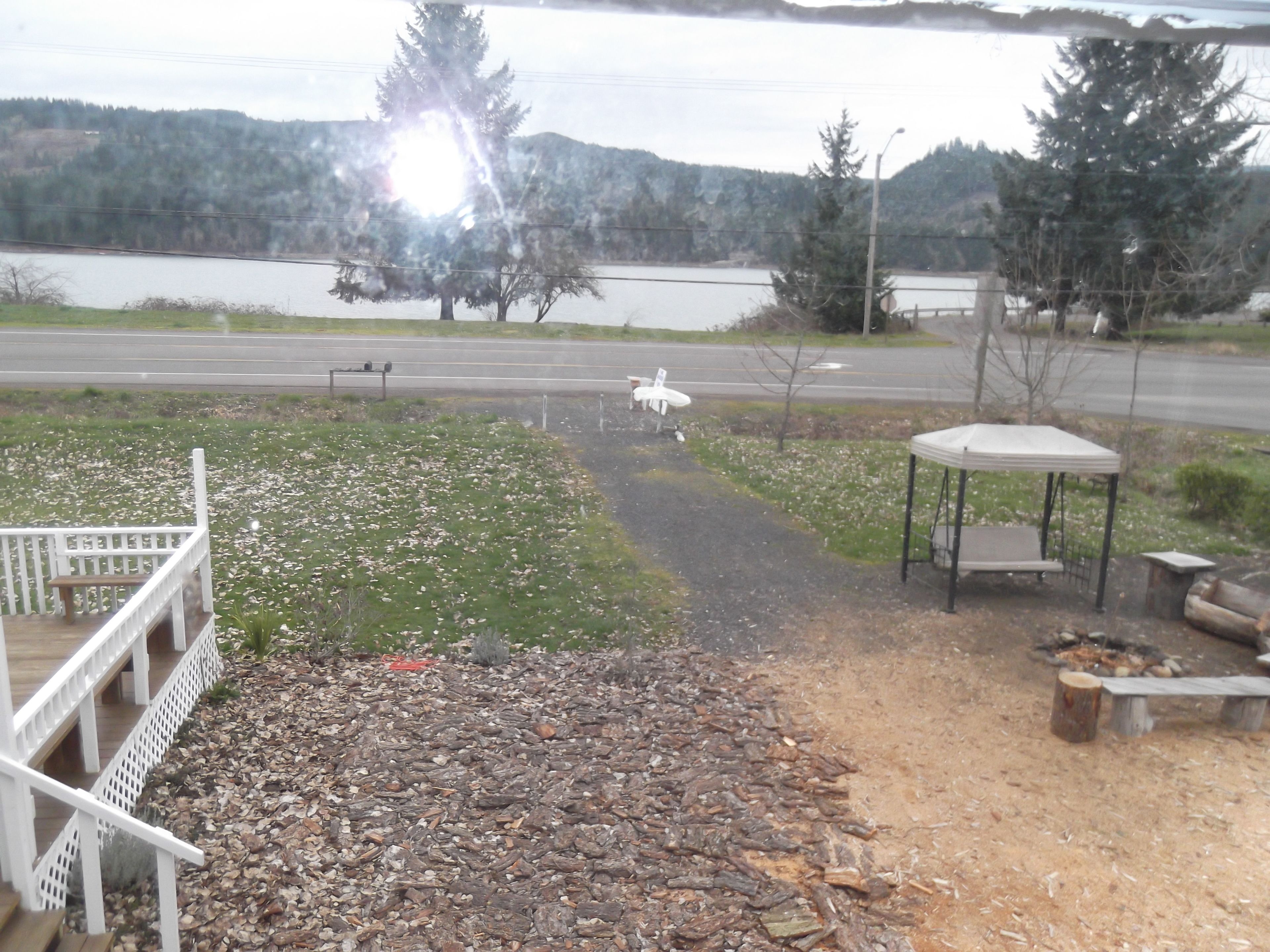 Vista al lago con una terraza de madera, una zona de estar cubierta y un camino de grava que lleva a un banco.