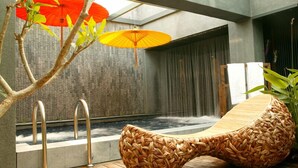 Zona d'una piscina interior moderna amb una cadira de relaxació de fusta, dues escales metàl·liques per a la piscina i una cascada.