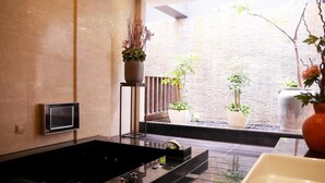 Un bany modern amb una banyera negra gran, un petit lavabo i un vàter. Hi ha diverses plantes en testos i un finestral gran que deixa entrar llum natural.