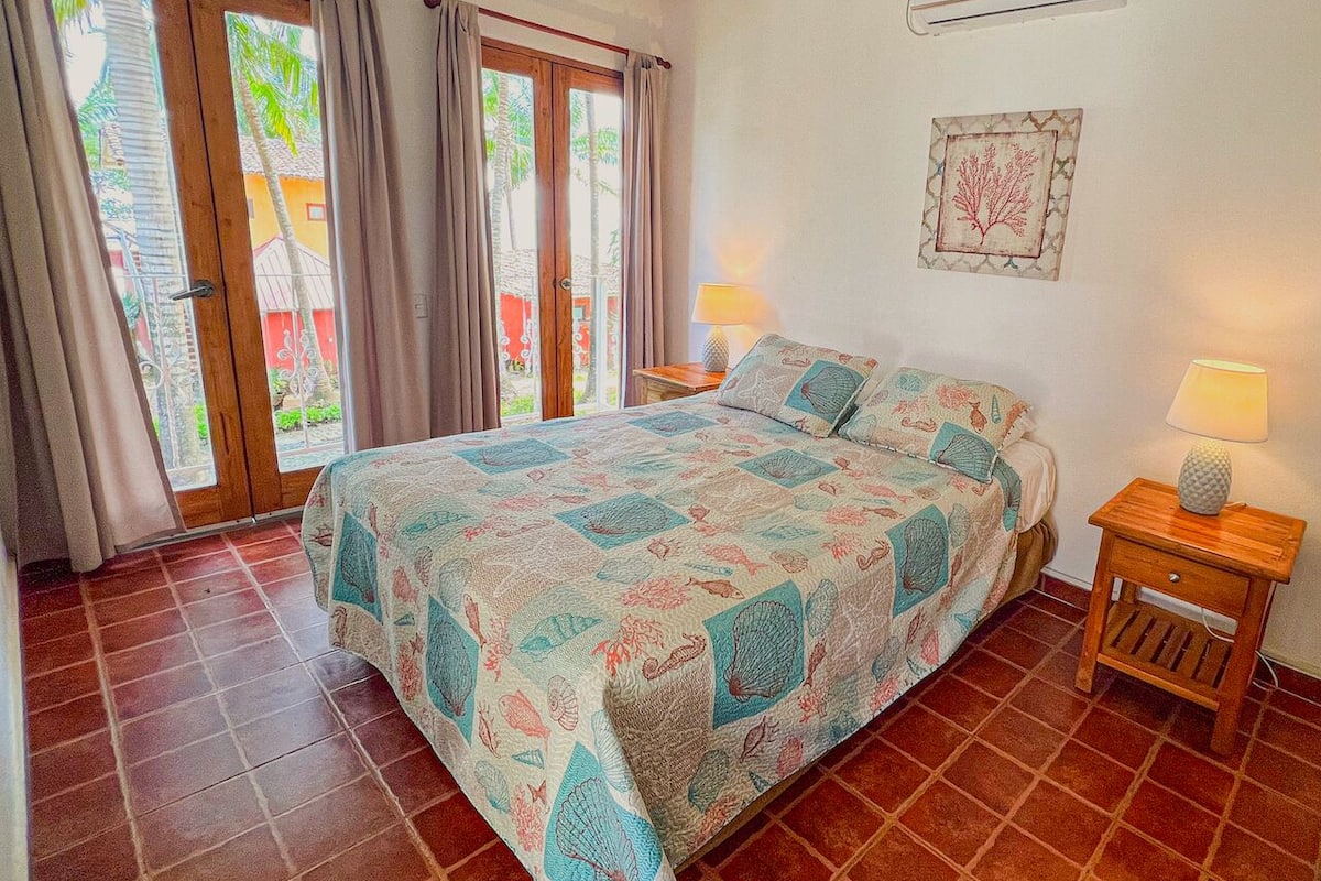 Un dormitorio con una cama, dos mesitas de noche, aire acondicionado y una ventana con cortinas.