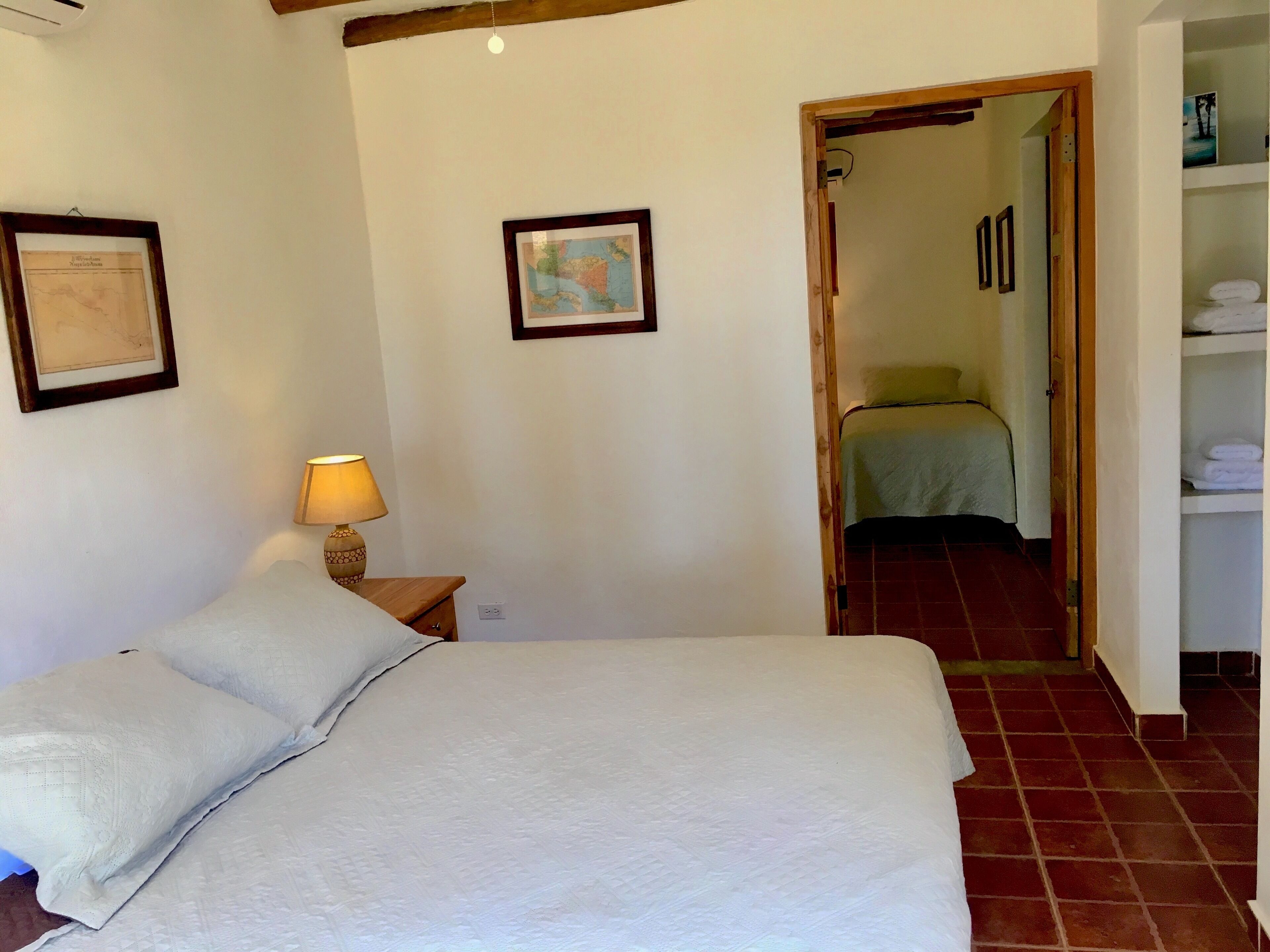 Un dormitorio con una cama, una mesita de noche con lámpara, un mapa en la pared y una puerta que da a otra habitación.