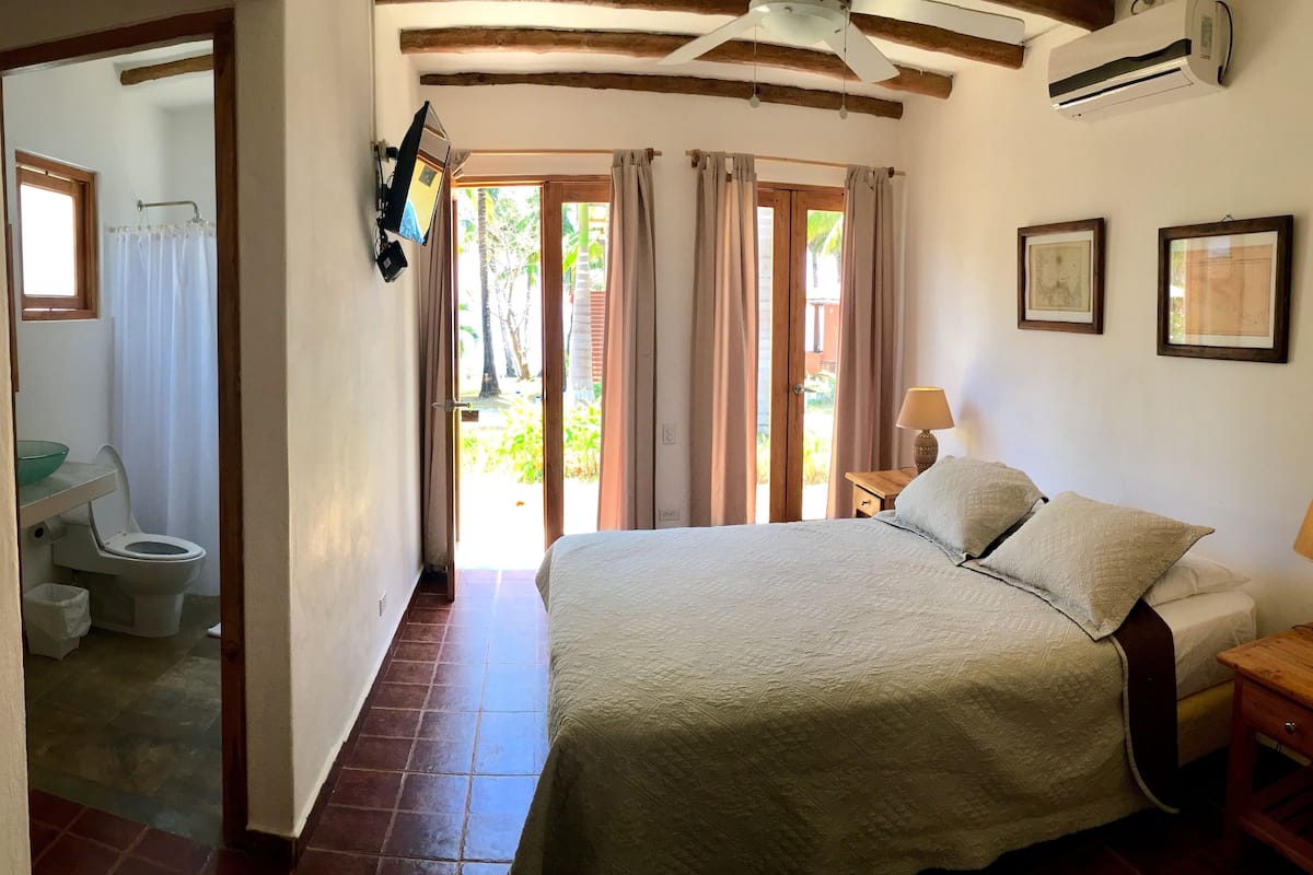 Un dormitorio con cama, mesita de noche, lámpara, ventana con cortinas y vistas al exterior.