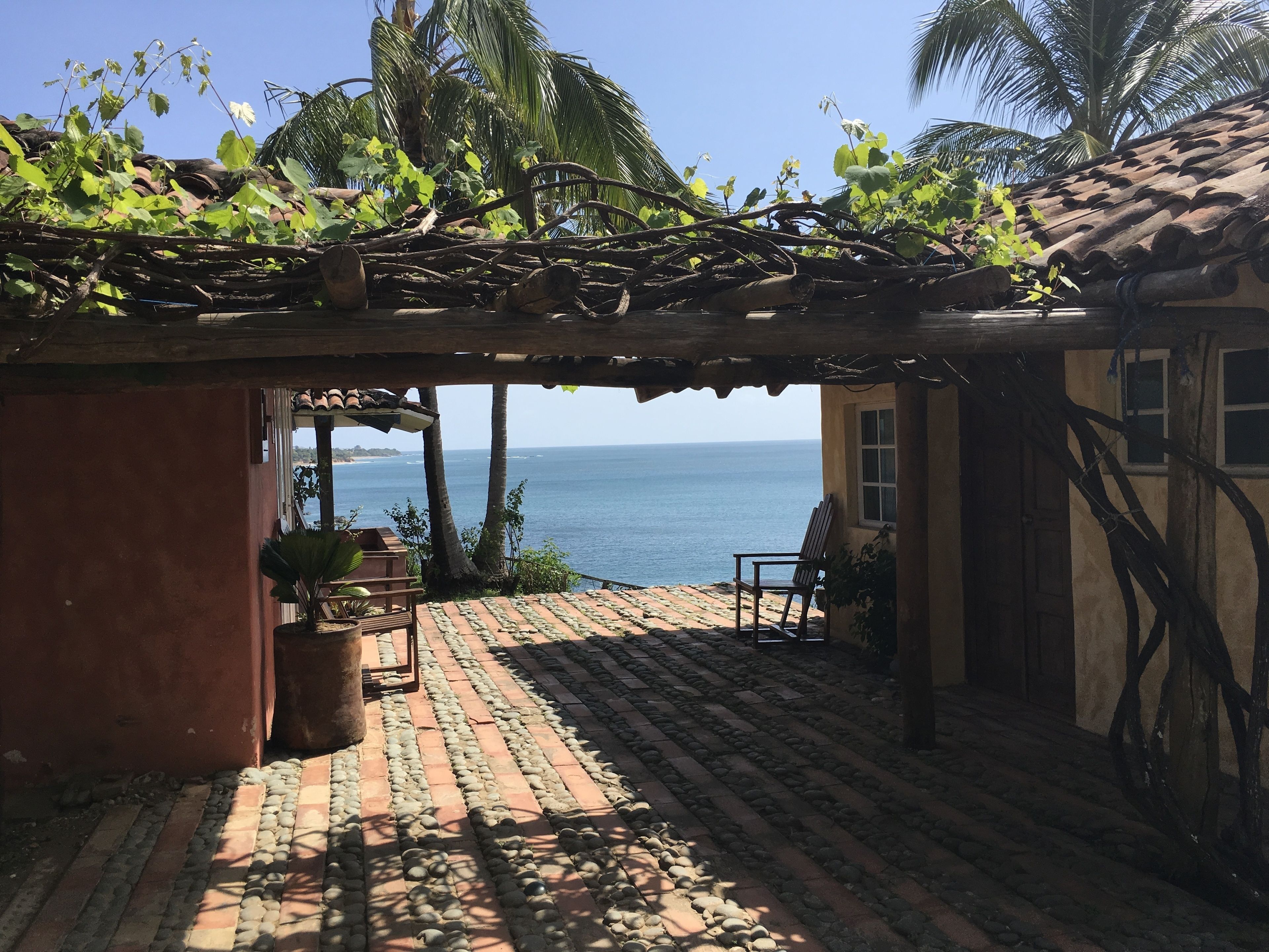 Un espacio al aire libre techado con vistas al océano, que presenta un suelo de baldosas, una pérgola de madera y una silla.