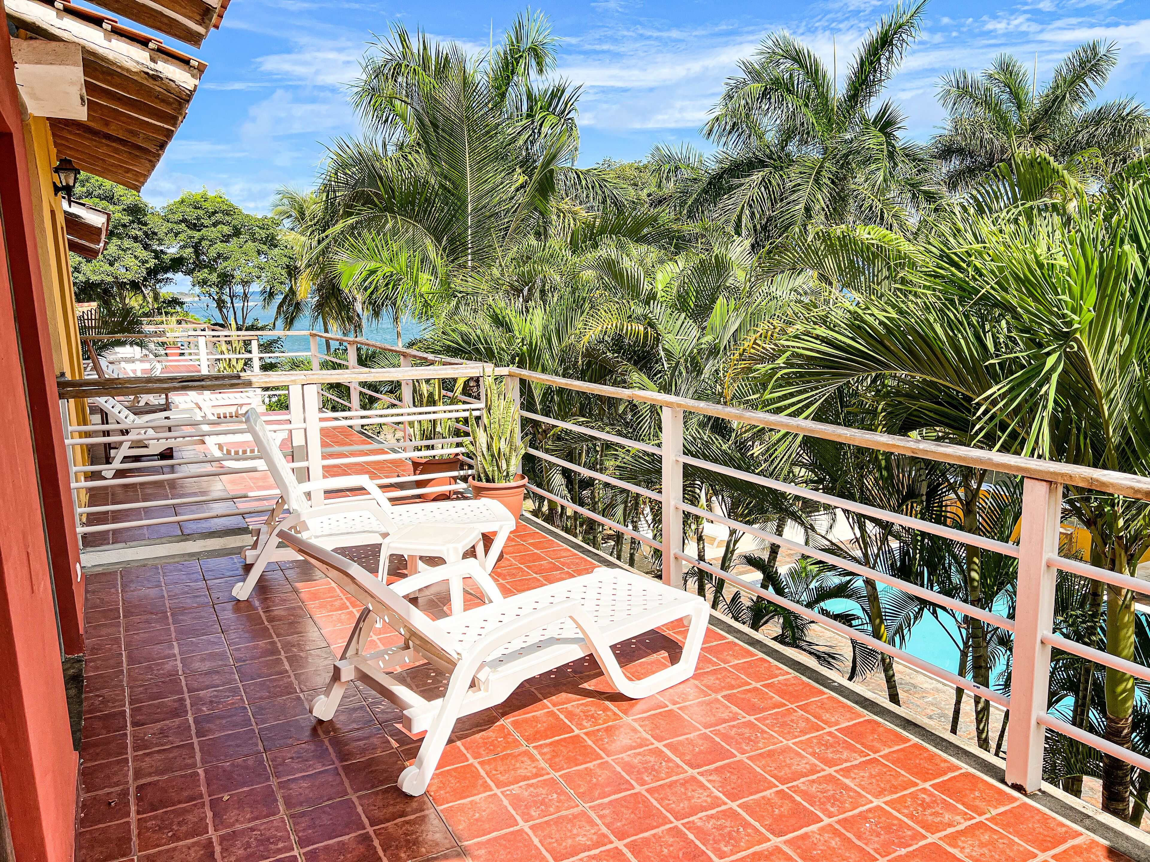 Un balcon avec des chaises en plastique blanc et un sol carrelé, donnant sur une piscine et une végétation luxuriante.