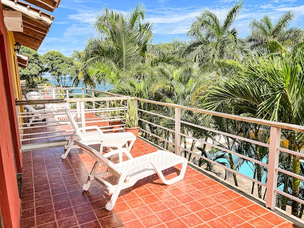 Een balkon met witte kunststof stoelen en een betegelde vloer, met uitzicht op een zwembad en weelderige begroeiing.