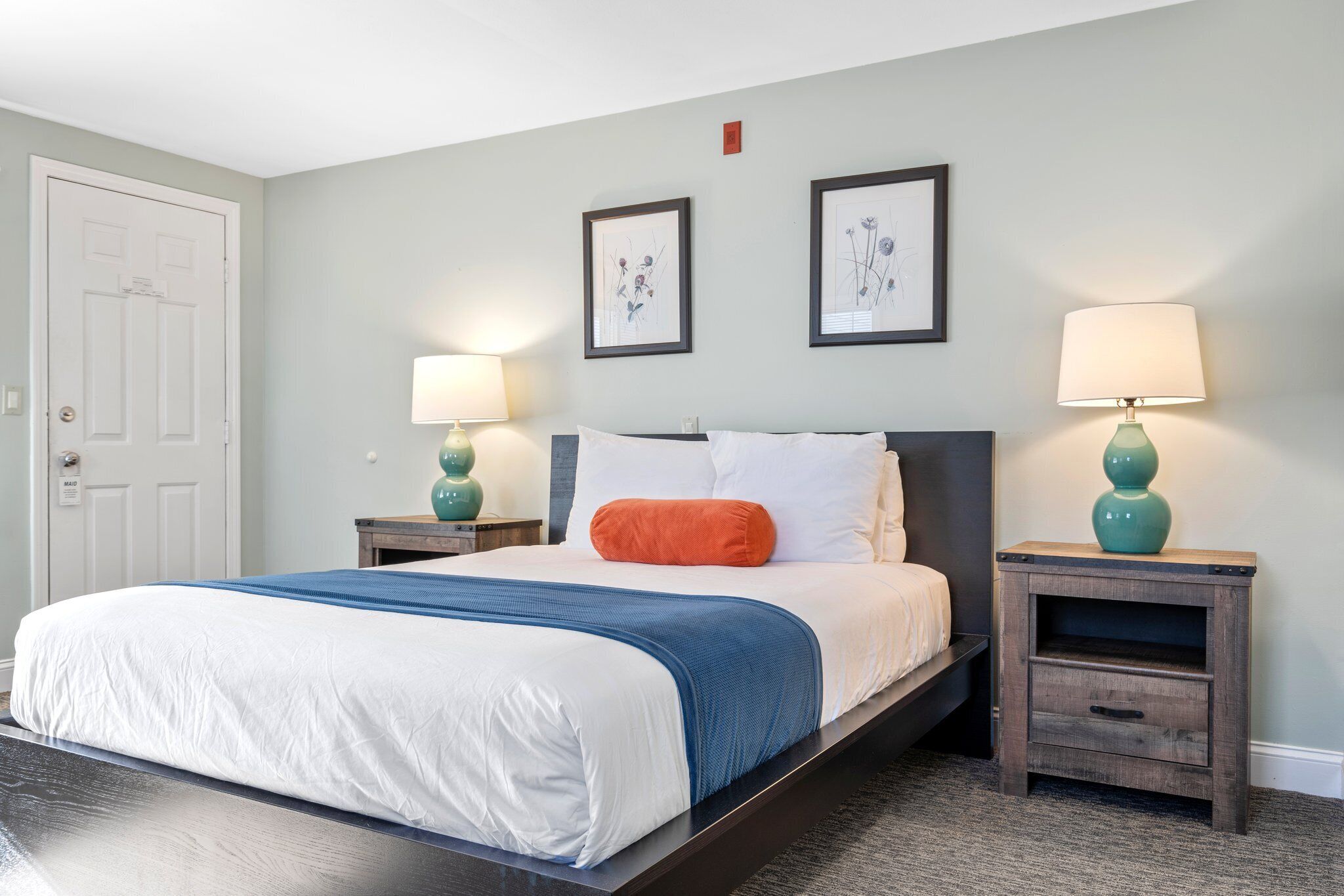 Une chambre à coucher comprenant un lit, deux tables de chevet, une commode, une porte et deux tableaux encadrés accrochés au mur.
