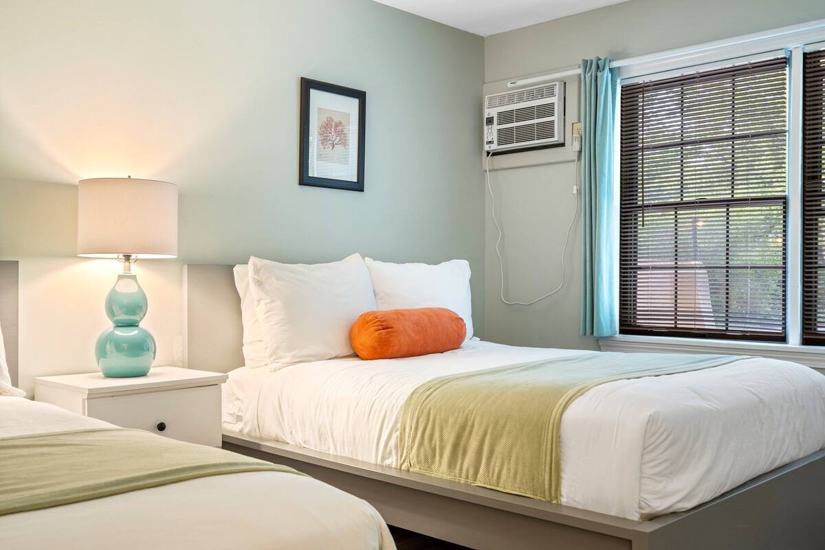 A bedroom with a bed, a nightstand with a lamp, an air conditioning unit, and a window with blinds.