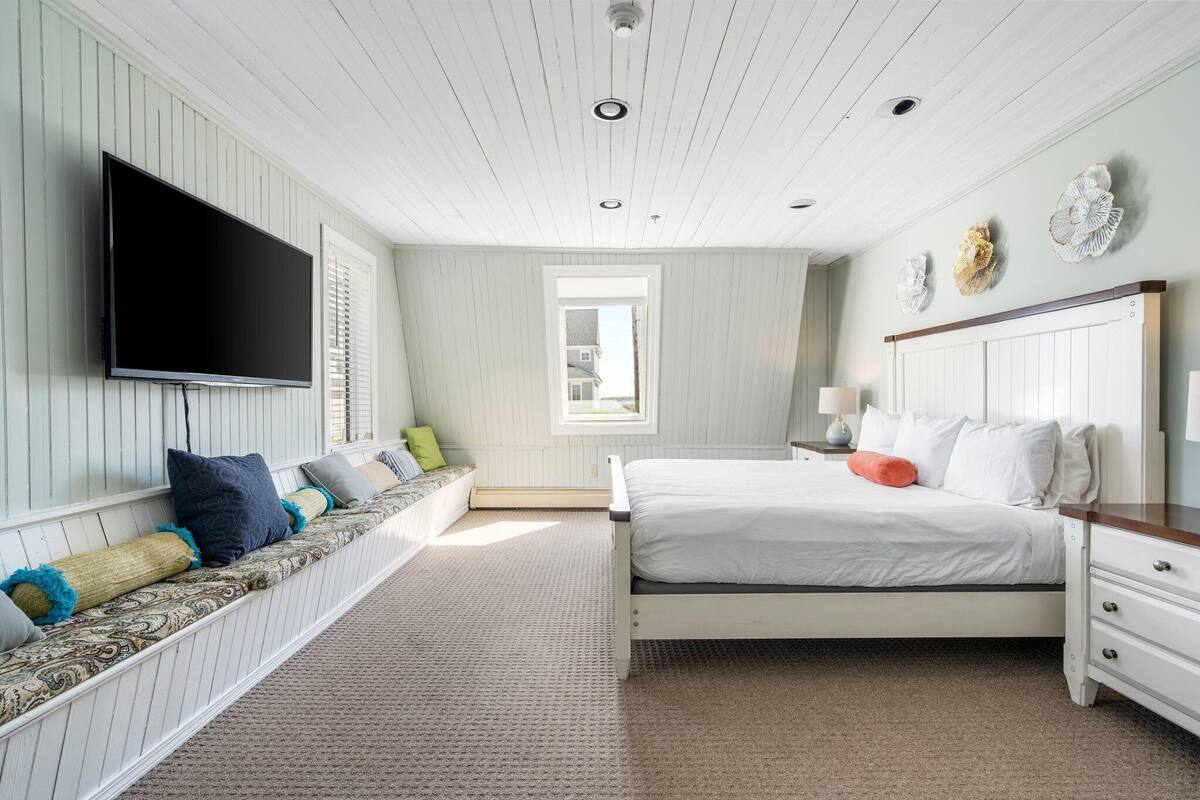 A bedroom with a large bed, a TV mounted on the wall, a window, and a bench with pillows.