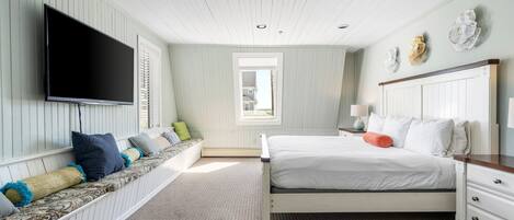 A bedroom with a large bed, a TV mounted on the wall, a window, and a bench with pillows.