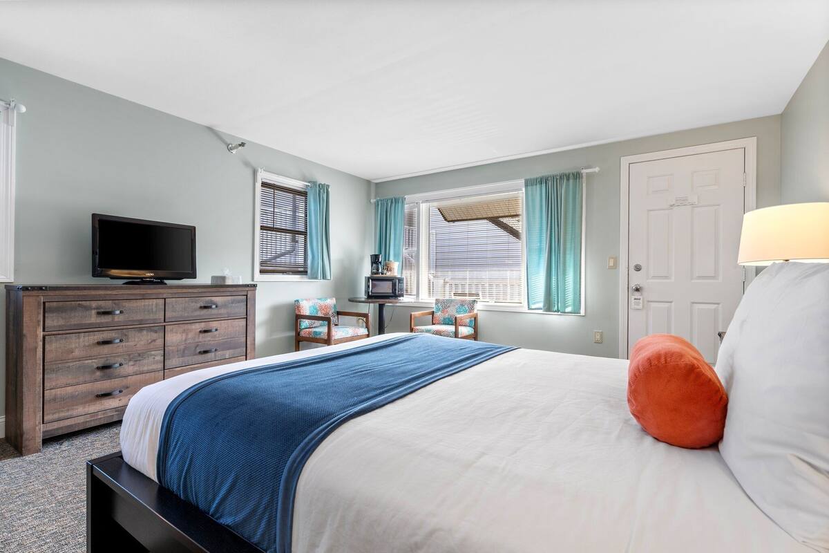 A bedroom with a bed, a dresser, a television, two chairs, and a window with curtains.