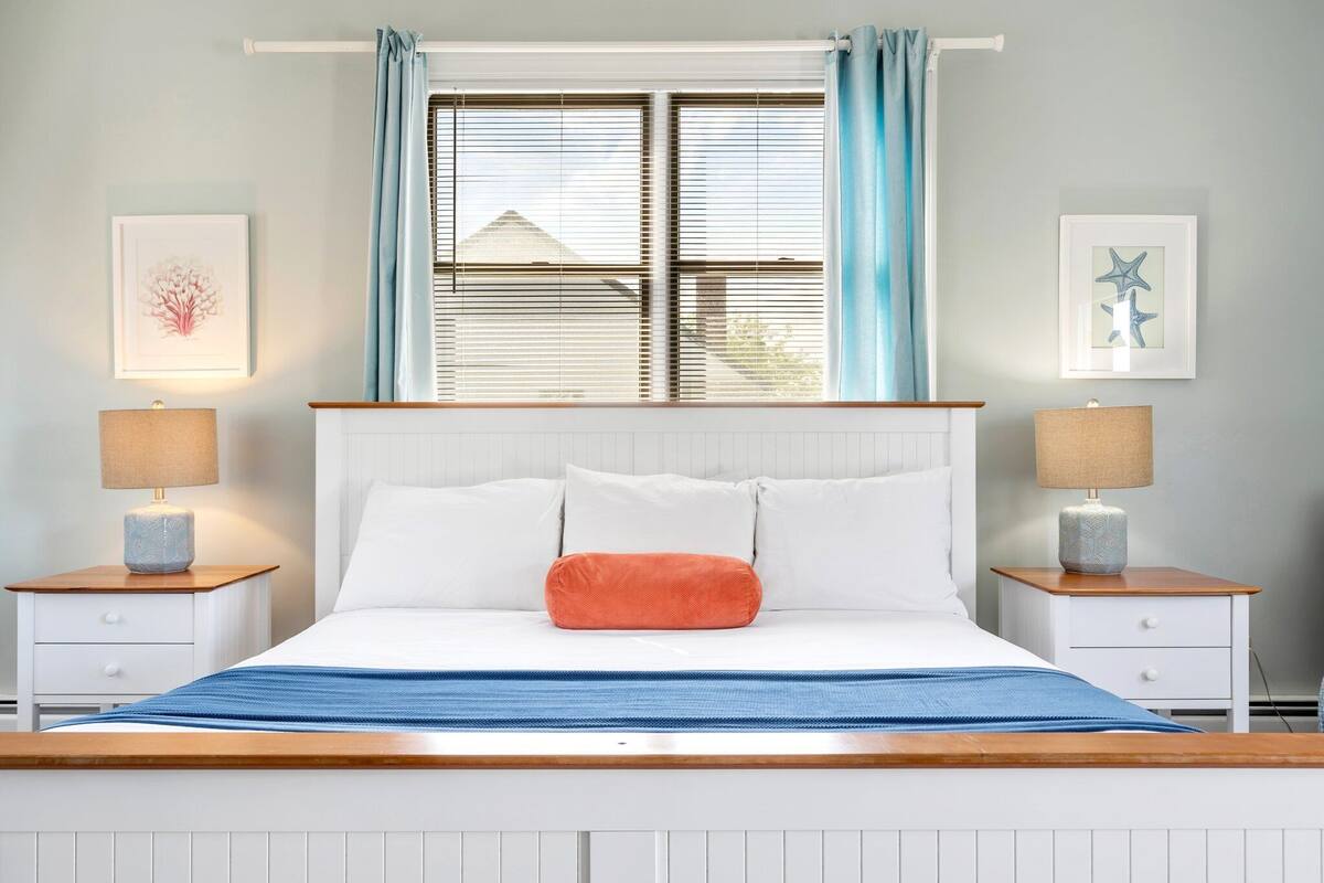 A bedroom with a bed, two bedside tables with lamps, a window with curtains, and two framed pictures on the wall.