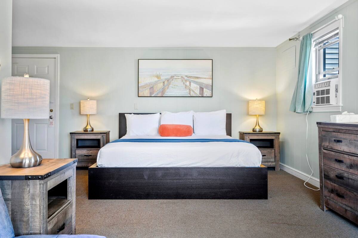 A bedroom with a large bed, two bedside tables with lamps, a wooden nightstand, a dresser, and a painting of a beach scene on the wall.