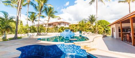 A swimming pool with a blue mosaic design, surrounded by palm trees and a building with a tiled roof.
