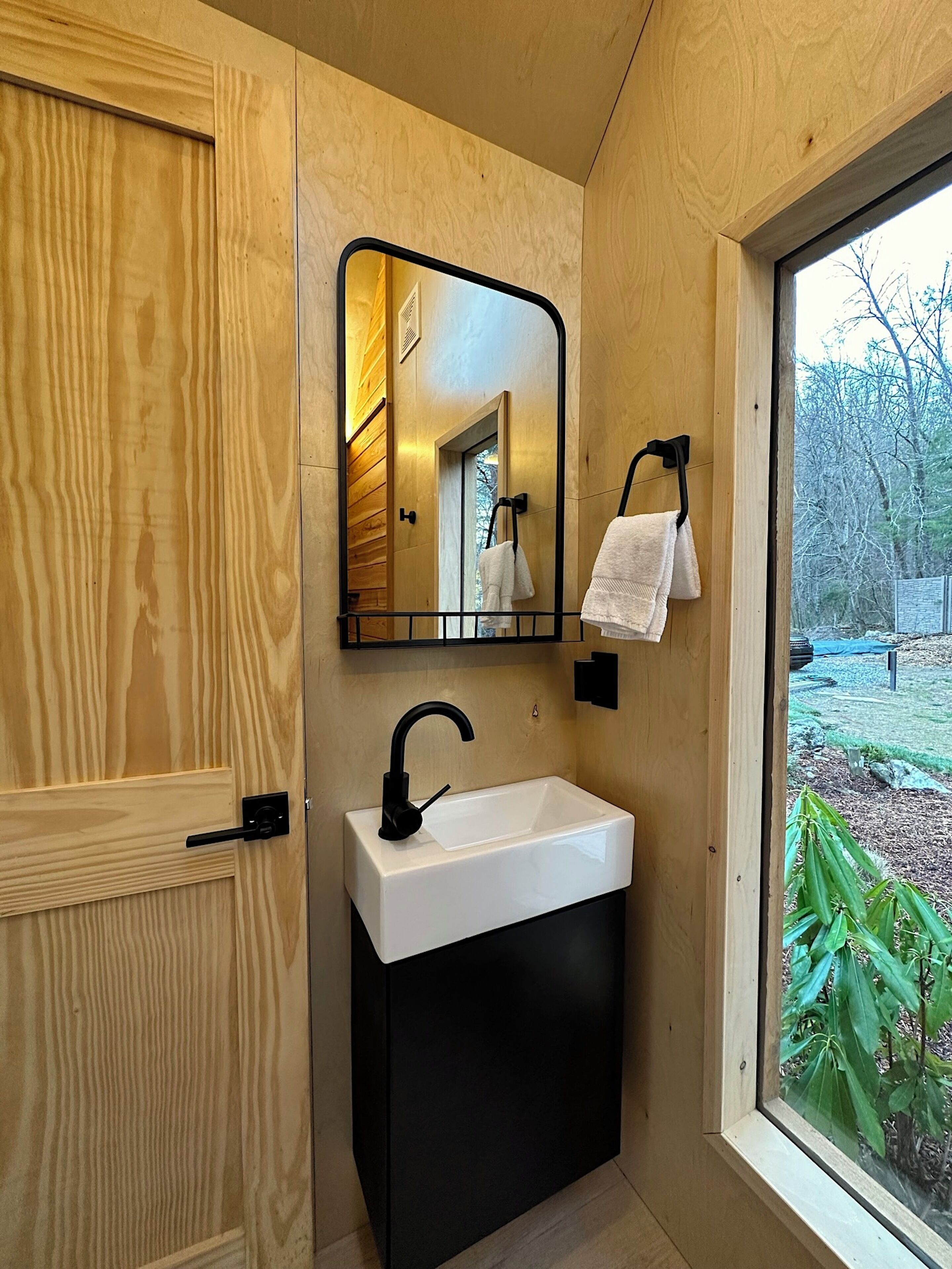 Une salle de bain moderne avec un meuble-lavabo en bois, un lavabo blanc, un robinet noir, un miroir et une vue sur l’extérieur par une fenêtre.