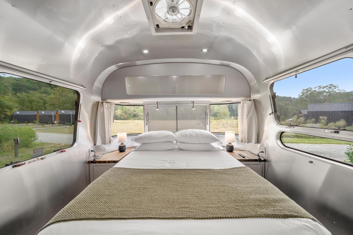 Ein Schlafzimmer mit Bett, Kissen, Nachttischen und einem Fenster mit Blick auf Grünflächen.