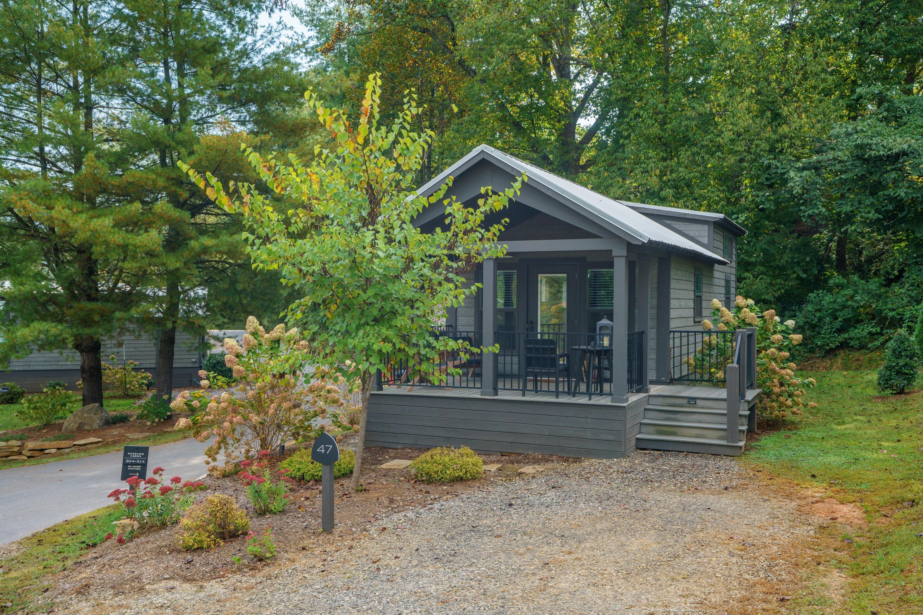 A small, single-story building with a porch and outdoor seating area, surrounded by trees and a gravel driveway.