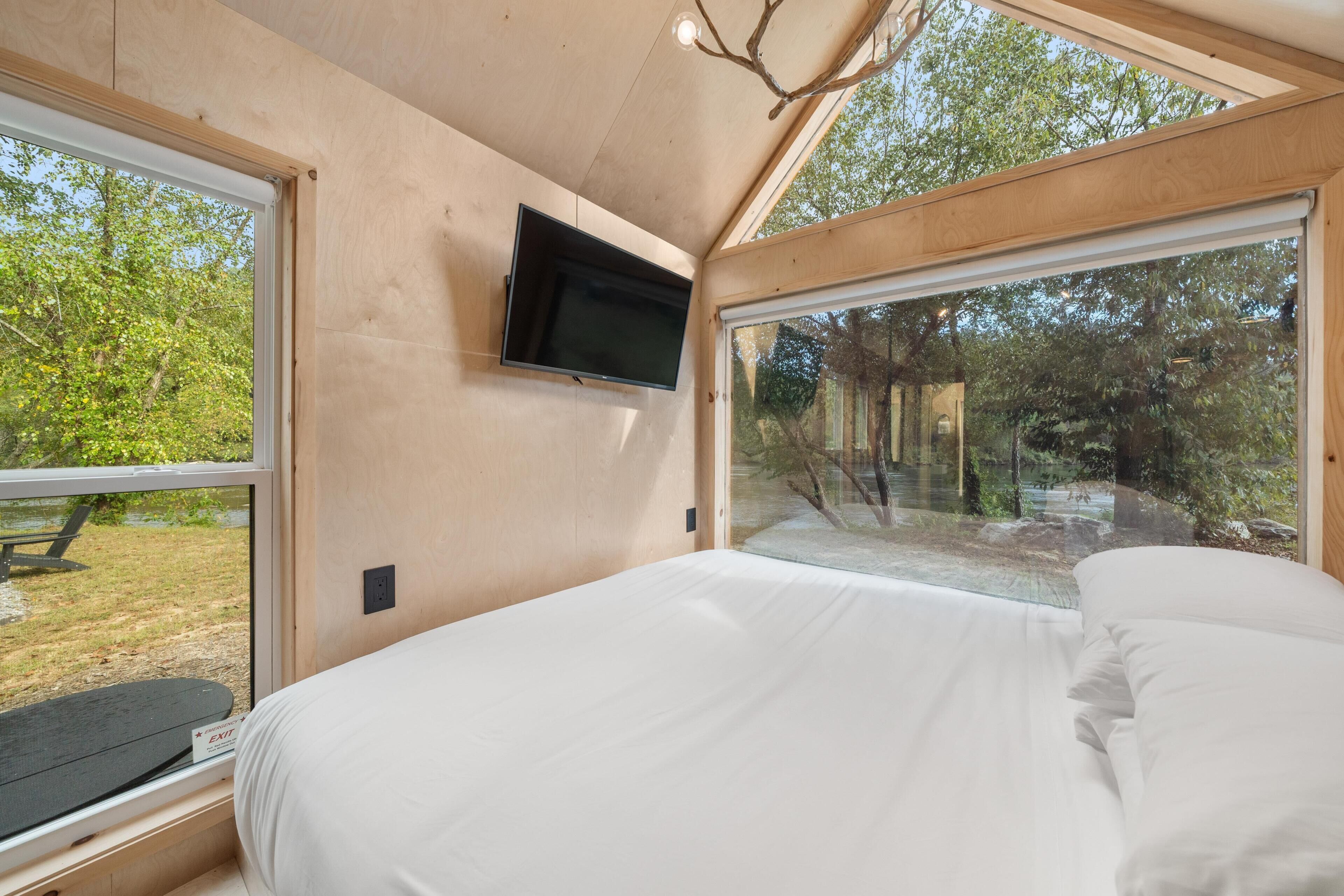 Ein Schlafzimmer mit einem Bett, einem an der Wand montierten Fernseher und großen Fenstern mit Blick ins Freie.