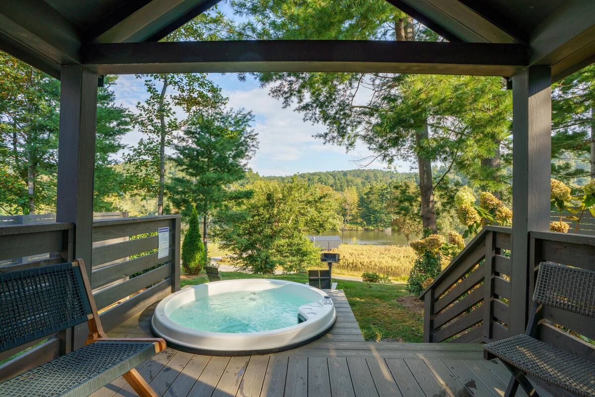 Un bain à remous sur une terrasse en bois, avec des chaises, donnant sur une forêt et un lac.