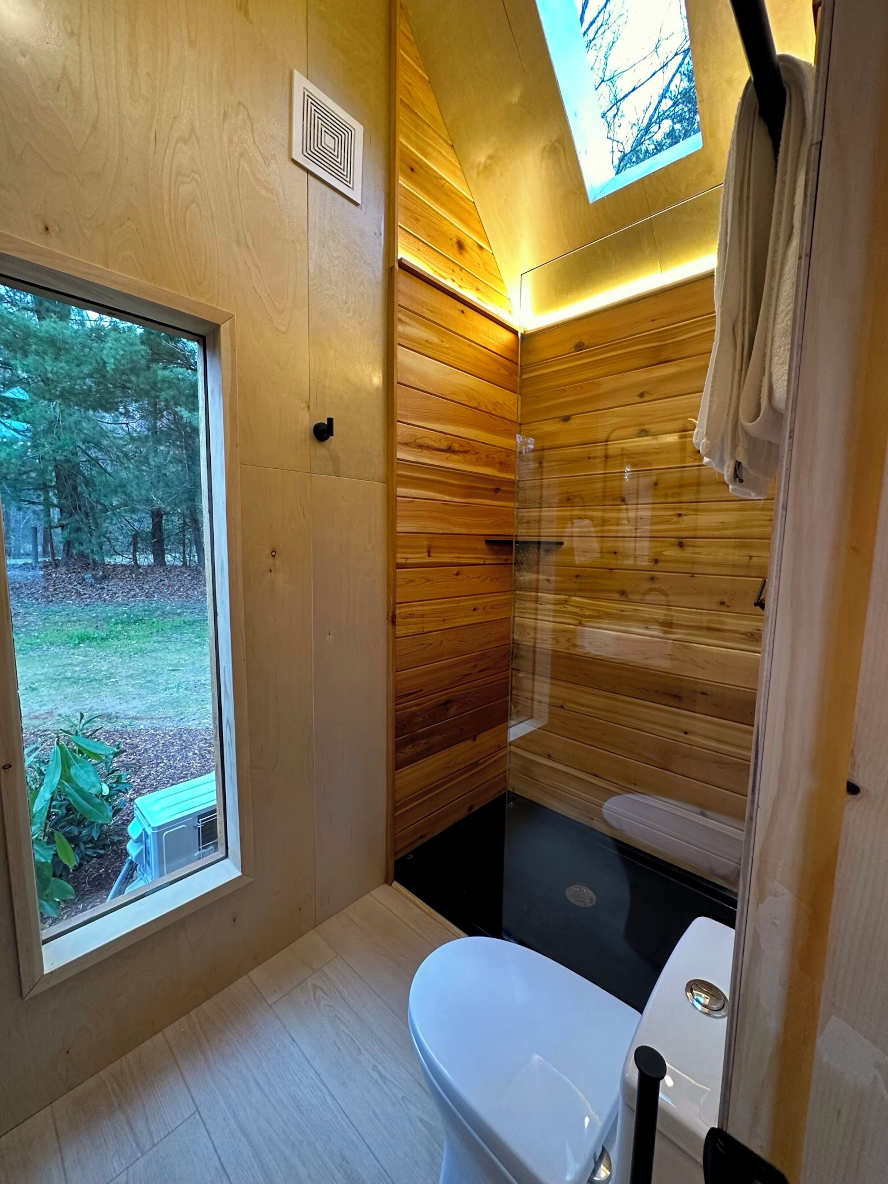 A compact wooden bathroom with a toilet, sink, and shower. A window offers a view of the outdoors.