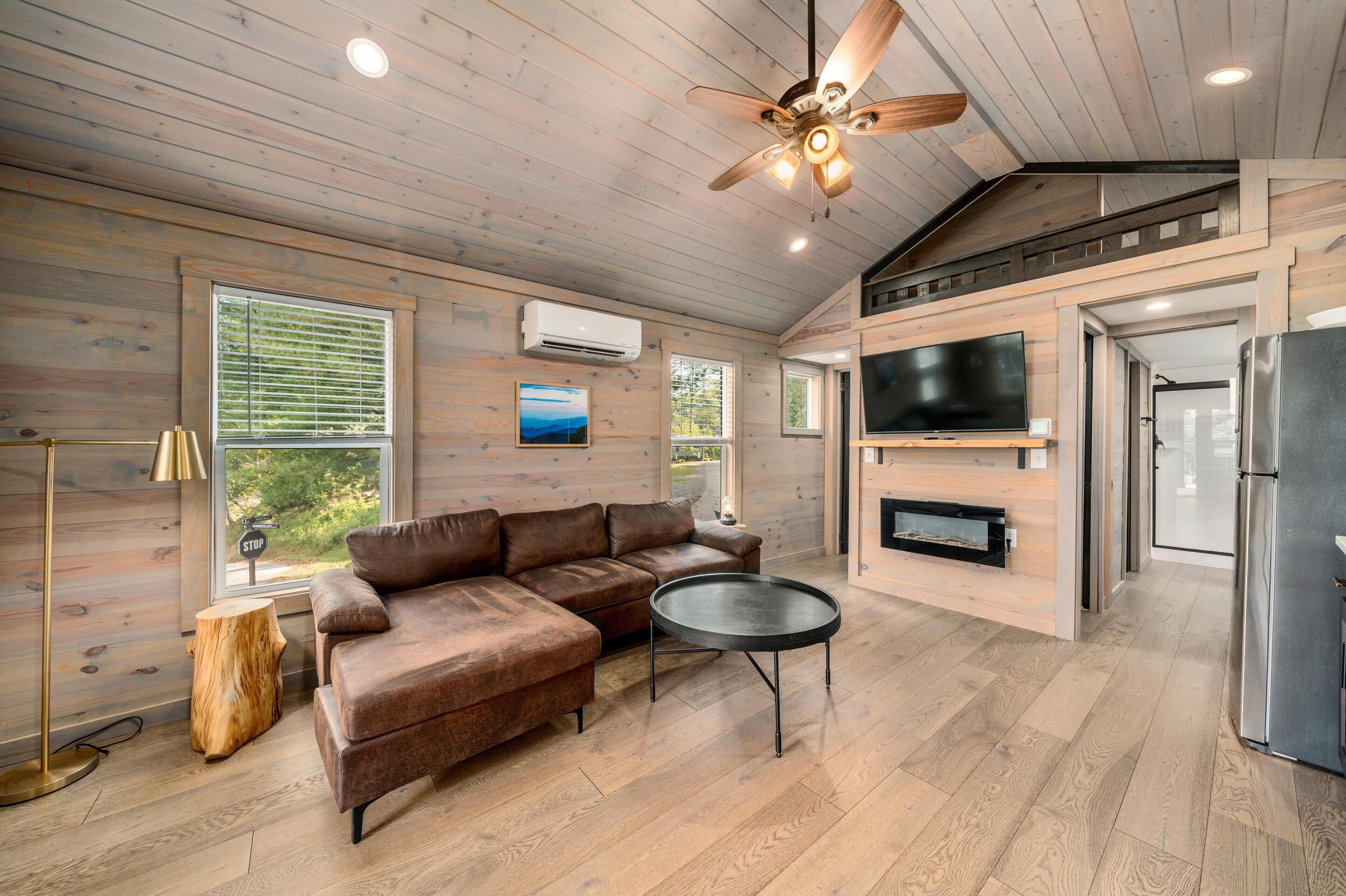 A modern cabin interior with a sectional sofa, a round coffee table, a ceiling fan, a TV, and a refrigerator.