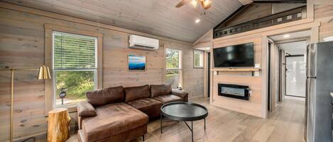 A modern cabin interior with a sectional sofa, a round coffee table, a ceiling fan, a TV, and a refrigerator.