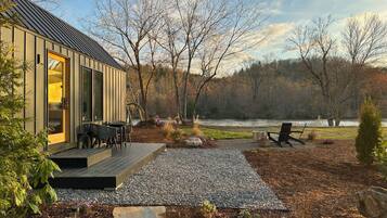A modern cabin with a deck, outdoor seating, and a view of a river and forest.