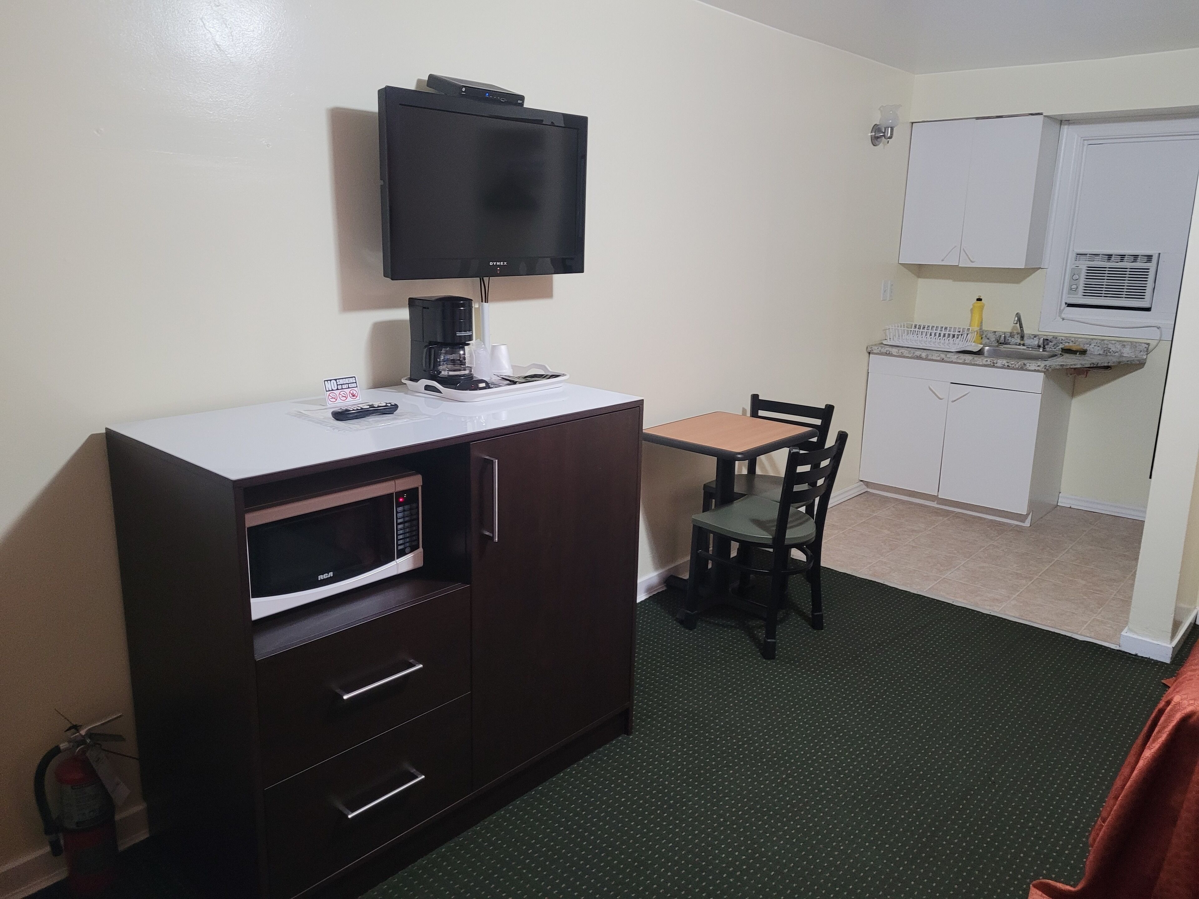 A compact hotel room with a kitchenette, a dining area with a table and chairs, a flat-screen TV mounted on the wall, and a microwave.