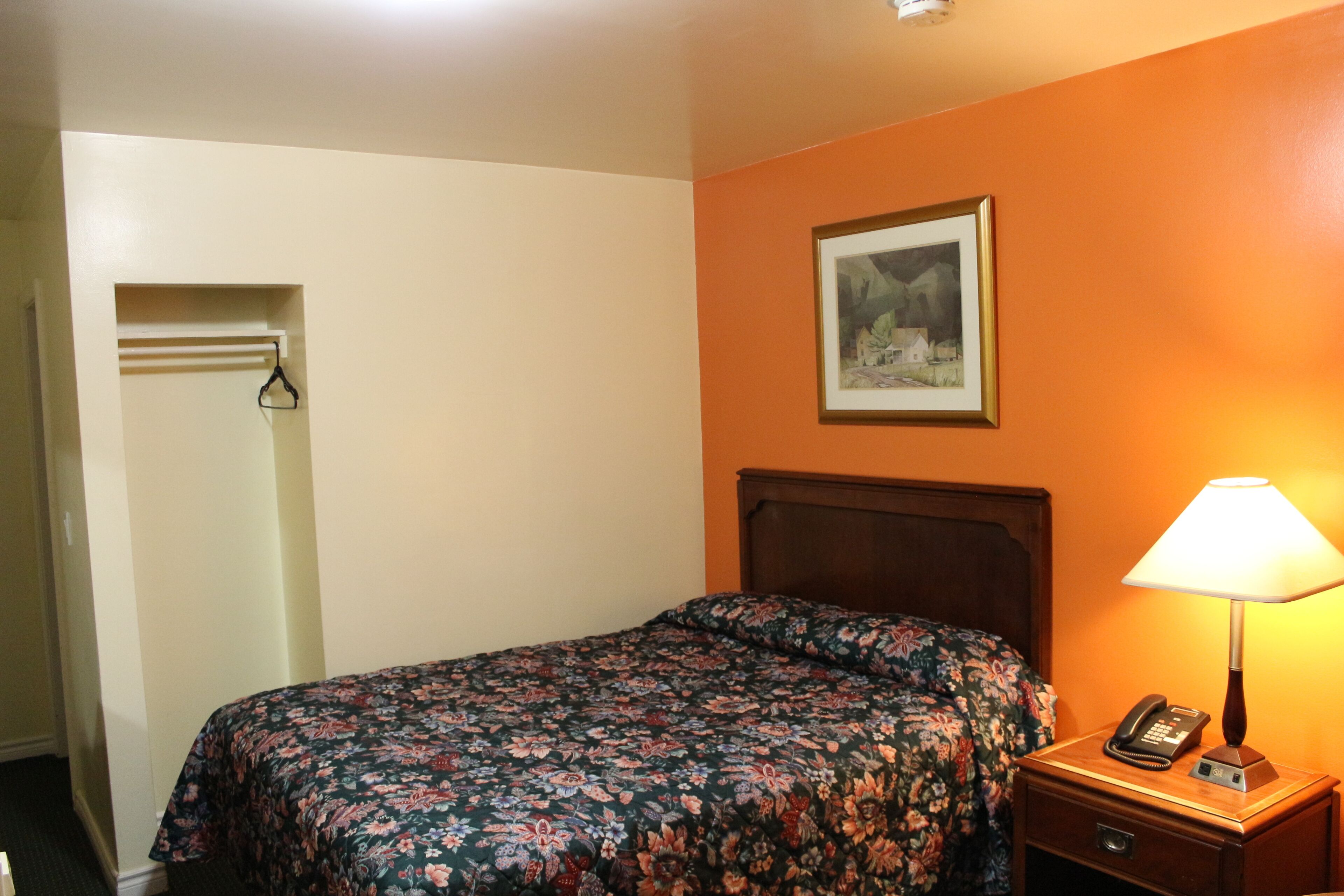 A hotel room with a bed, a nightstand with a lamp, a framed picture on the wall, and a door with a coat rack.