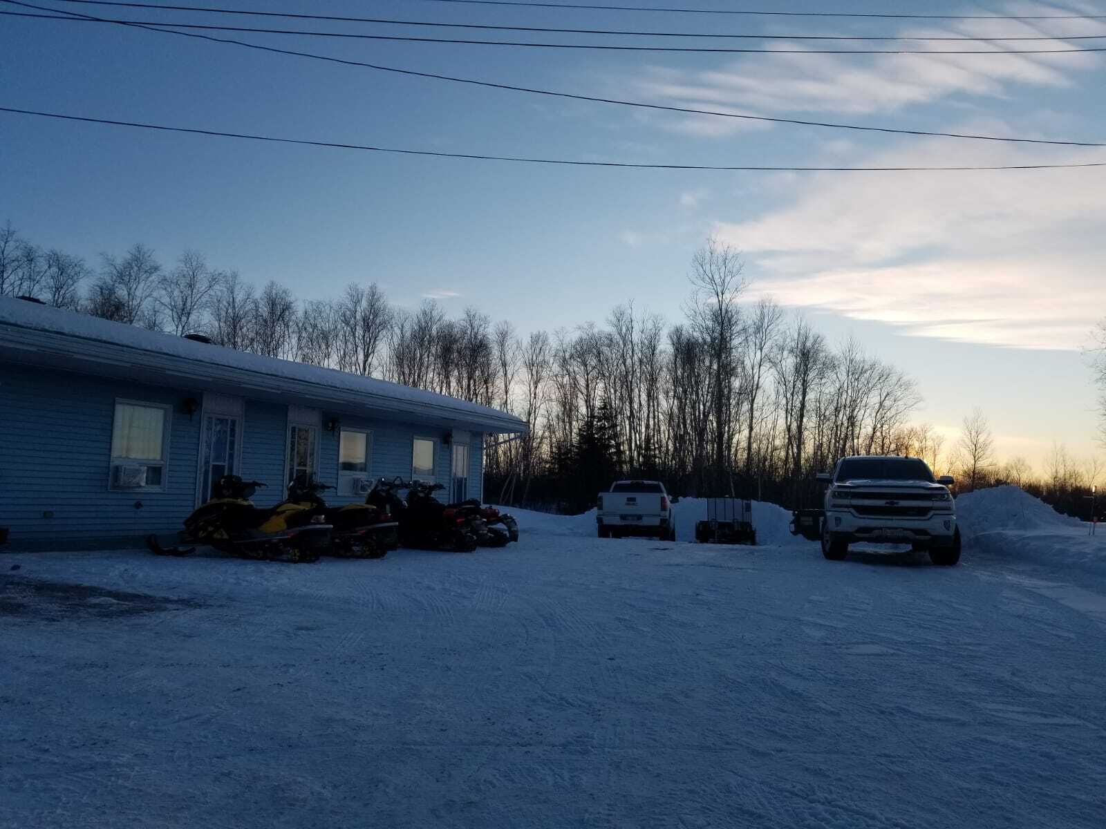 Un estacionamiento cubierto de nieve con un edificio azul, varias motocicletas todo terreno y un SUV blanco.